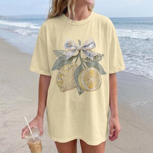 Peut inclure: T-shirt jaune clair avec un motif de citron. Le motif comprend un nœud, des citrons et des feuilles. Les citrons sont jaunes et blancs, avec un motif à carreaux. Les feuilles sont vertes et grises. Le nœud est bleu et blanc. Le t-shirt est à manches courtes et col rond.