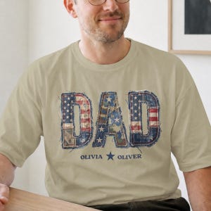 以下が含まれることがあります： ベージュ色のTシャツ。アメリカ国旗の模様をあしらったパッチワークデザインで「DAD」の文字が描かれています。「DAD」の下には「OLIVIA」と「OLIVER」の名前が星で区切られています。
