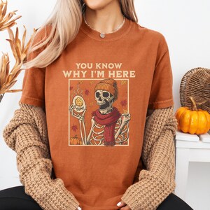 Peut inclure: T-shirt orange brûlé avec un motif de squelette portant un bonnet et une écharpe, tenant un biscuit et une tasse. Le texte "YOU KNOW WHY I'M HERE" est au-dessus du motif. Fait partie d'une collection sur le thème de l'automne.