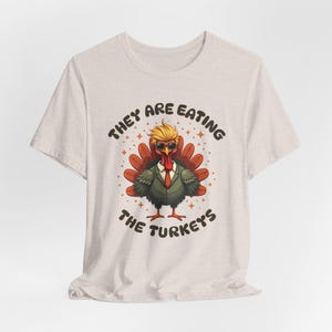 Puede incluir: Una camiseta beige con un pavo de dibujos animados que lleva gafas de sol y un traje. El texto "They are eating the turkeys" está impreso en la camiseta.