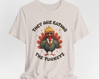 Może przedstawiać: Beżowa koszulka z kreskówkowym indykiem w okularach przeciwsłonecznych i garniturze. Na koszulce nadrukowany jest tekst "They are eating the turkeys".