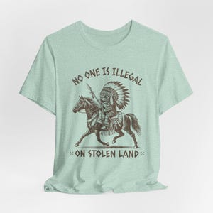 May include: A light green t-shirt featuring a graphic of a Native American riding a horse. The graphic is in brown and black and the text on the shirt reads "No one is illegal on stolen land."