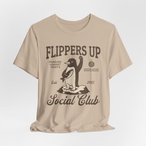 May include: A tan t-shirt with a distressed graphic featuring a penguin holding a fish. The text reads "FLIPPERS UP" above "PENGUINS AGAINST TARIFFS" and "SOCIAL CLUB". Additional text includes "Beard Island and McDonald Islands 2025" and "Est. 2025".