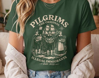 Op de afbeelding: Een donkergroen T-shirt met korte mouwen en een crèmekleurige afbeelding van drie figuren in historische kleding, een zeilschip en de tekst: PILGRIMS THE ORIGINAL ILLEGAL IMMIGRANTS Estd. 1620. Het ontwerp bevat een kompas en een perkamentrol.