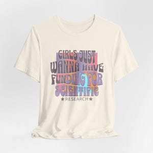 Pode incluir: Uma camiseta de cor creme com uma estampa gráfica que diz "Girls Just Wanna Have Funding For Scientific Research" em uma fonte retrô com blocos coloridos.