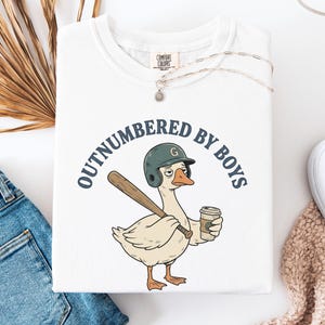 May include: White t-shirt with a graphic of a goose wearing a baseball helmet and holding a bat and coffee cup. The text "OUTNUMBERED BY BOYS" is printed above the goose. A silver necklace is draped over the shirt.