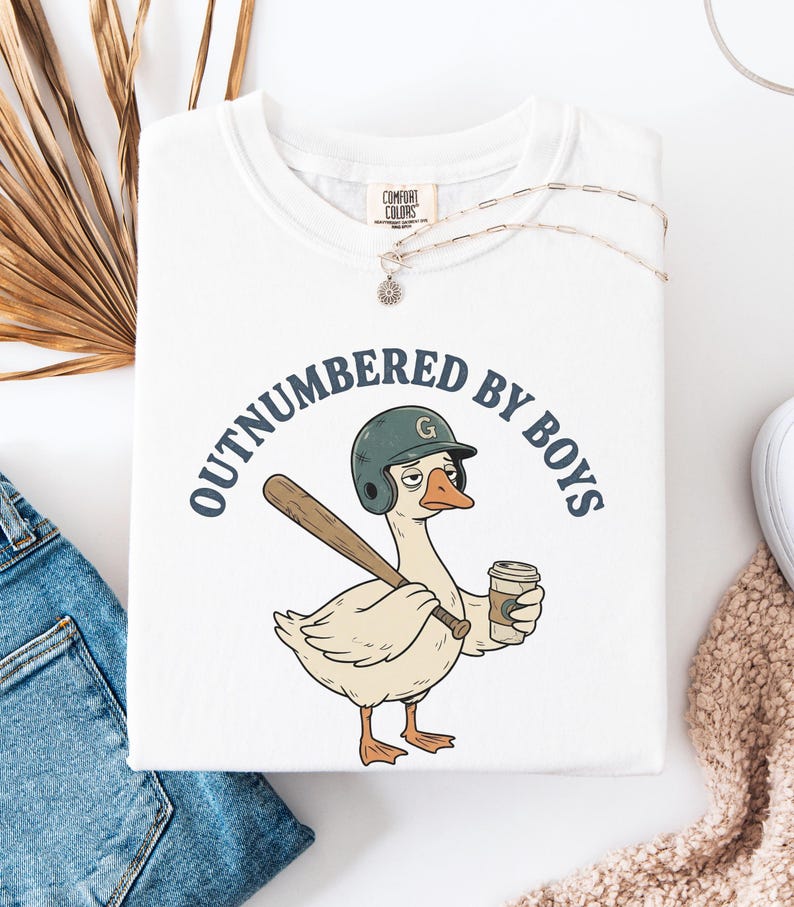 May include: White t-shirt with a graphic of a goose wearing a baseball helmet and holding a bat and coffee cup. The text "OUTNUMBERED BY BOYS" is printed above the goose. A silver necklace is draped over the shirt.