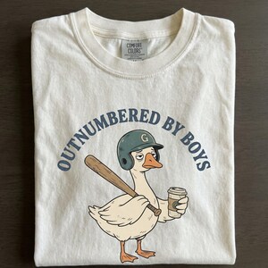 May include: Cream-colored t-shirt with the phrase "OUTNUMBERED BY BOYS" in navy blue. A cartoon goose wearing a baseball helmet and holding a bat and coffee cup is featured in the center. The shirt is folded on a dark surface.