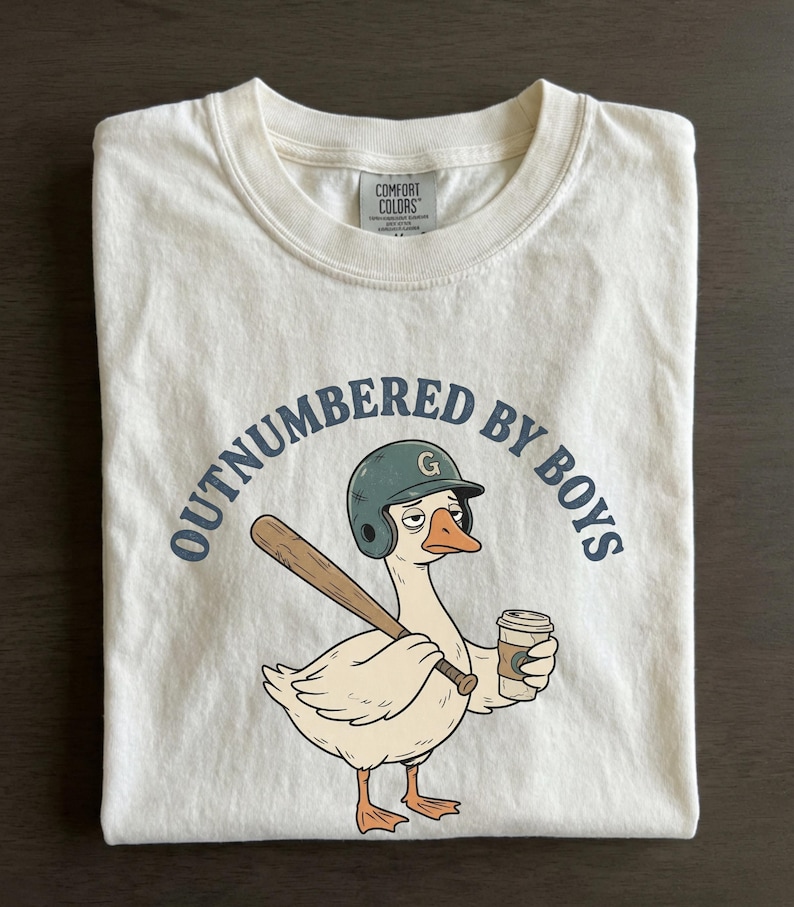 May include: Cream-colored t-shirt with the phrase "OUTNUMBERED BY BOYS" in navy blue. A cartoon goose wearing a baseball helmet and holding a bat and coffee cup is featured in the center. The shirt is folded on a dark surface.