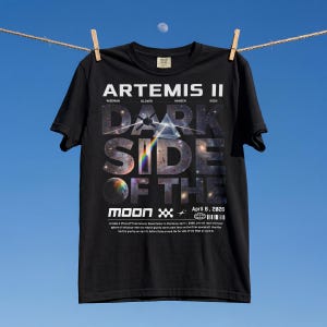 May include: Black t-shirt with the text "ARTEMIS II DARK SIDE OF THE MOON" in white and gray. The design includes a space-themed graphic with a rainbow prism and a planet. The date "April 6, 2026" is also printed on the shirt.