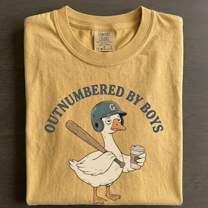 May include: Mustard yellow t-shirt with the phrase "OUTNUMBERED BY BOYS" in navy blue. The graphic features a cartoon goose wearing a baseball helmet and holding a bat and a coffee cup. The shirt is folded.