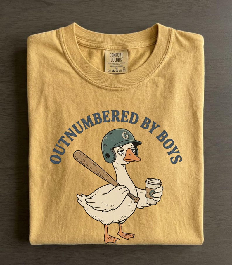 May include: Mustard yellow t-shirt with the phrase "OUTNUMBERED BY BOYS" in navy blue. The graphic features a cartoon goose wearing a baseball helmet and holding a bat and a coffee cup. The shirt is folded.