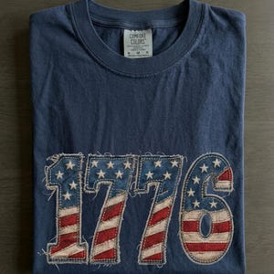 May include: Navy blue t-shirt with the number "1776" appliqued on the front. The numbers are designed with a distressed American flag pattern, featuring red and white stripes and white stars on a blue background.