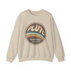 Könnte beinhalten: Beigefarbenes Sweatshirt mit Rundhalsausschnitt und einem kreisförmigen Aufdruck. Der Aufdruck zeigt das Wort "PLUTO" in Retro-Schrift, mit bunten Streifen und dem Text "NEVER FORGET 1930-2006". Das Sweatshirt hat lange Ärmel und eine klassische Passform.