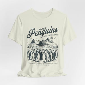 Könnte beinhalten: Ein weißes T-Shirt mit einer schwarz-weißen Grafik von Pinguinen in einer schneebedeckten Landschaft. Der Text "Make Penguins Tax-Free Again" ist auf dem Shirt aufgedruckt. Die Koordinaten "53.1060 S, 73.5042 E" und der Text "Heard Island and McDonald Islands" sind ebenfalls auf dem Shirt aufgedruckt.