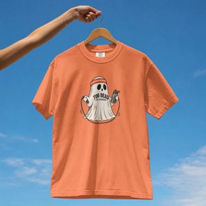 Może przedstawiać: Pomarańczowy t-shirt z krótkim rękawem z grafiką białego ducha w pomarańczowej opasce na głowie, trzymającego skakankę i butelkę z wodą. Napis "TOO DEAD TO CARDIO" jest nadrukowany czarnymi literami na środku klatki piersiowej ducha.