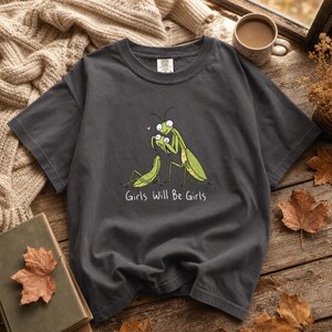 Peut inclure: Un t-shirt gris anthracite à manches courtes avec une illustration graphique de deux mantes religieuses vertes, l'une debout et l'autre assise, avec un petit cœur rose au-dessus et le texte blanc Girls Will Be Girls imprimé en dessous.