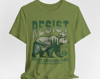 Könnte beinhalten: Grünes T-Shirt mit einem Aufdruck eines Bären mit Rangerhut und dem Text "Resist" und "Protect our National Parks - Parks over Profit -"