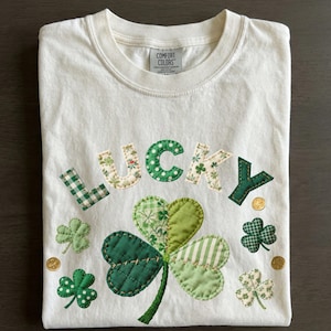 Puede incluir: Camiseta color crema con la palabra "LUCKY" aplicada en varias telas verdes y estampadas. Un gran trébol y tréboles más pequeños están debajo de la palabra, junto con monedas de oro. La camiseta está doblada cuidadosamente.