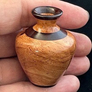 May include: A small, handcrafted wooden vase with a dark brown rim and base, and a contrasting dark brown band around the middle. The vase has a light brown, swirling wood grain pattern. The vase is approximately 2.5 inches tall.