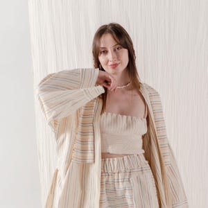 May include: A woman wearing a striped, cream-coloured cardigan over a white ribbed tank top and matching striped trousers. She is standing in front of a white textured background.