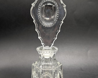 May include: A clear glass perfume bottle with a hexagonal base and a tall, ornate stopper. The bottle features embossed oval panels surrounded by beaded detailing on each side. The stopper has a matching beaded oval design at the top.