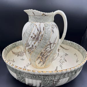 May include: A vintage porcelain wash basin set featuring a pitcher and bowl. The set has a cream and green color scheme with floral and geometric patterns. The pitcher has a handle and a spout, and the bowl has a wide rim.