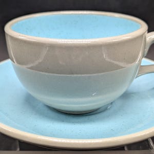 May include: A ceramic teacup and saucer set. The cup and saucer are light blue with a speckled texture, and the rim is a contrasting beige color. The interior of the cup is a brighter blue. The cup has a curved handle.