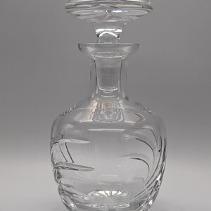 May include: A clear glass decanter with a stopper. The decanter has a round body with a textured design and a tall, slender neck. The stopper is also clear glass and has a round, flat top.