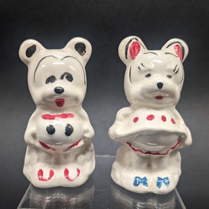 May include: Two white ceramic salt and pepper shakers shaped like bears. The bear on the left has black eyes and a red mouth. The bear on the right has black eyes and a red mouth with a red and white polka dot dress.