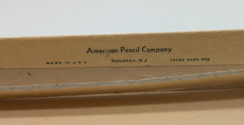 May include: Close-up of a beige box with black text that reads "American Pencil Company, Hoboken, N.J., Trade Mark Reg., Made in U.S.A."