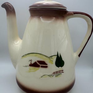 May include: A white ceramic coffee pot with a brown lid and handle. The pot has a brown and green painted scene of a house, trees, and a fence.