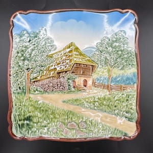 May include: Square ceramic plate with a raised relief design depicting a rural scene. The image features a stone house with a thatched roof, a dirt path, trees, and a blue sky. The plate has a brown border and is primarily green, brown, and blue.