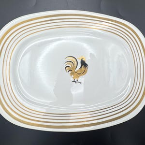 May include: White oval serving platter with gold stripes around the rim. A gold, black, and brown rooster is centered on the platter. The platter is a decorative piece for serving food.