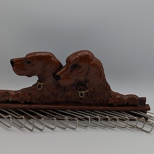 May include: A brown wooden tie rack featuring two sculpted dogs with gold-colored collars. The rack has a series of metal hooks for hanging ties. The dogs are lying down, and the rack is on a white surface.