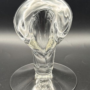 May include: A clear, elegant glass vase with a unique, curved design. The vase features a flared top and a slender stem that sits on a round base. The glass is transparent, allowing light to pass through, and is ideal for displaying flowers or as a decorative piece.