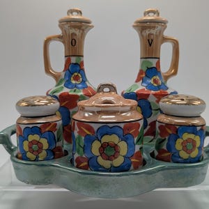 May include: A vintage ceramic condiment set featuring two oil and vinegar cruets, two salt and pepper shakers, and a lidded sugar bowl. The set is decorated with a floral pattern in blue, red, and yellow, and sits on a light green tray.