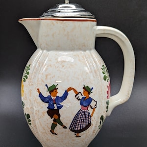 May include: A white ceramic pitcher with a silver lid and a painted scene of two people dancing. The pitcher has a brown rim and a floral design.