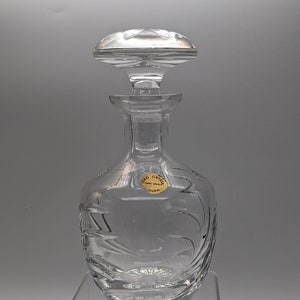 May include: A clear glass decanter with a stopper. The decanter has a decorative etched design and a label that reads "Lead Crystal Hand Made Portugal".