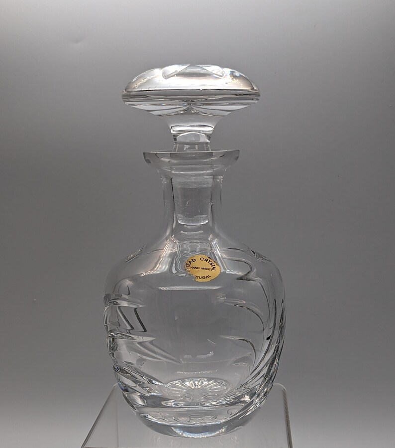 May include: A clear glass decanter with a stopper. The decanter has a decorative etched design and a label that reads "Lead Crystal Hand Made Portugal".