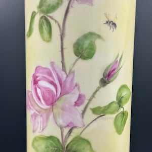 May include: A yellow ceramic vase with a floral design. The vase features pink roses with green leaves and two bees. The vase is decorated with a hand-painted design.