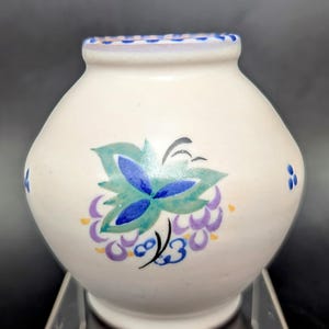 May include: A white ceramic vase with a floral design. The vase has a round body and a small opening at the top. The floral design is painted in shades of blue, green, purple, and yellow.