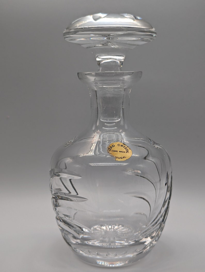 May include: A clear glass decanter with a stopper. The decanter has a swirl design and a gold label that reads "Lead Crystal Hand Made Portugal".