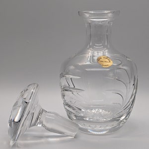 May include: A clear glass decanter with a textured design and a matching stopper. The decanter has a label that reads "Cristal Hand Made Portugal".