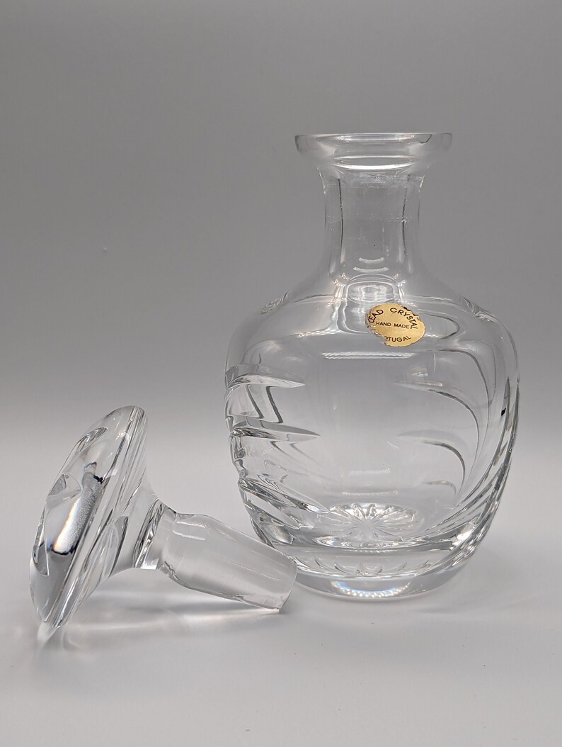 May include: A clear glass decanter with a textured design and a matching stopper. The decanter has a label that reads "Cristal Hand Made Portugal".