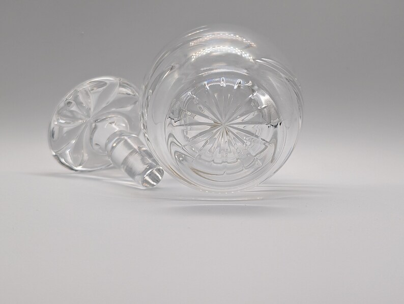May include: A clear glass bottle stopper with a decorative, starburst-shaped design on the bottom. The stopper has a round, flat top with a small, clear glass knob.