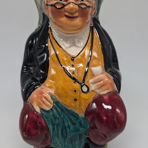 May include: A ceramic figurine of a man wearing a black hat, yellow vest, and red pants. He is holding a green cloth in his hand and has a white collar and a black chain around his neck.
