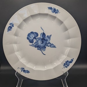 May include: White ceramic plate with a scalloped edge and a blue floral design. The plate features a central bouquet and smaller floral accents around the rim. The plate is displayed on a clear acrylic stand.