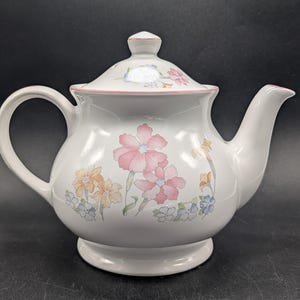 May include: White ceramic teapot with a lid, handle, and spout. The teapot is decorated with pink, yellow, and blue floral designs. The lid and rim have a pink trim. The teapot sits on a small, round base.