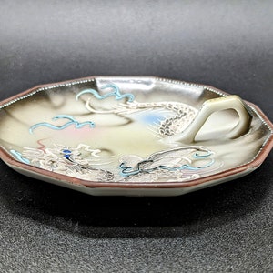 May include: A white porcelain dish with a dragon design in blue, pink, and gold. The dish has a handle and is octagonal in shape.